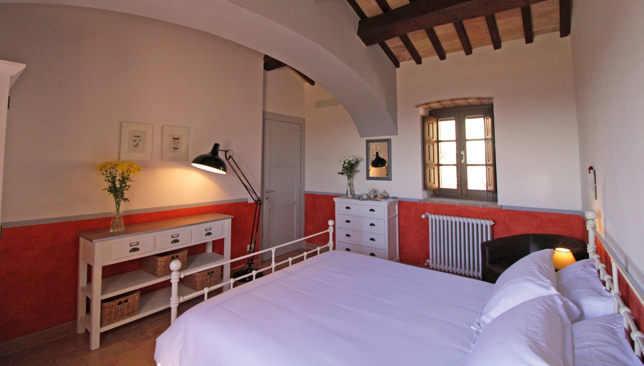 Vaulted bedroom with terracotta-red wall band, iron bed and a shuttered window