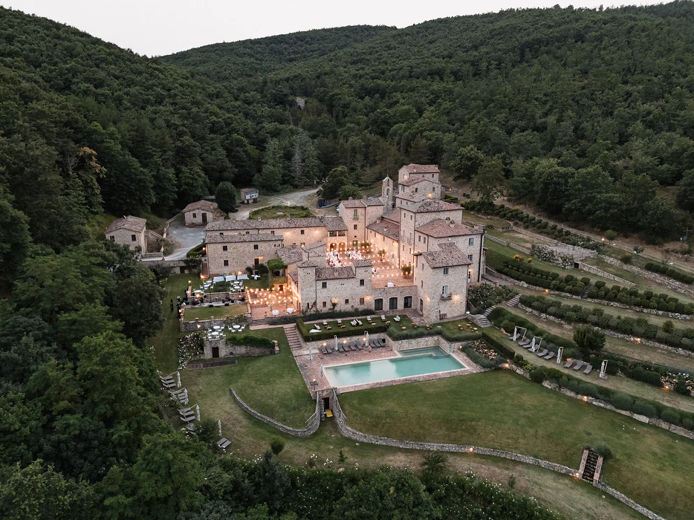 Aerial view at dusk of the restored medieval hamlet, its lit courtyard, pool and terraced gardens