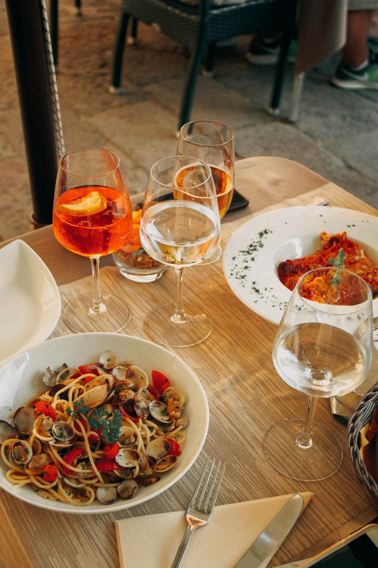 An outdoor table set with clam pasta, an Aperol spritz and glasses of chilled white wine