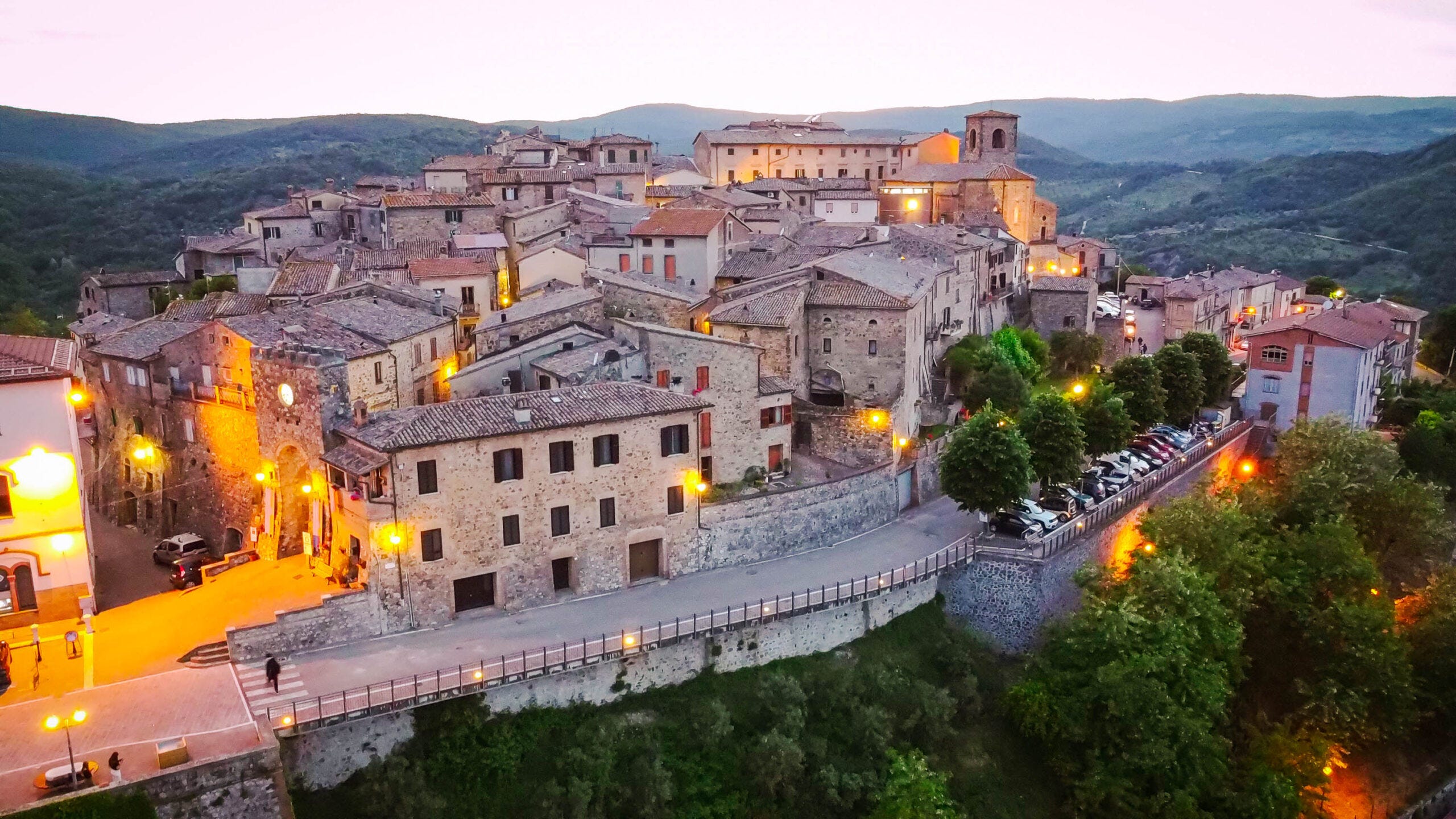 Dusk aerial panorama of Allerona, its stone houses and bell tower glowing on a hilltop above wooded valleys